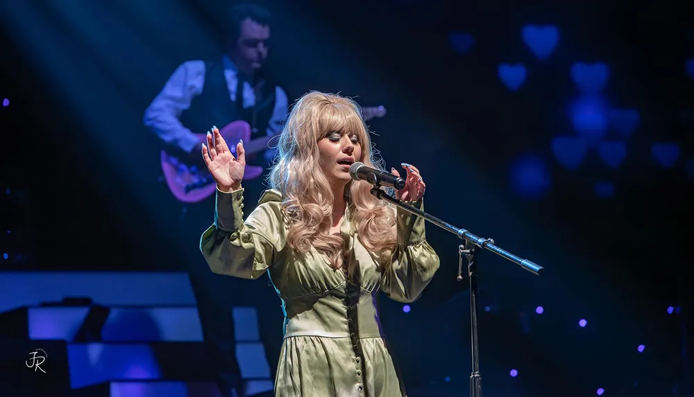 A woman with long blonde hair sings passionately into a microphone on stage accompanied by a guitarist in the background with heart-shaped lights illuminating the scene