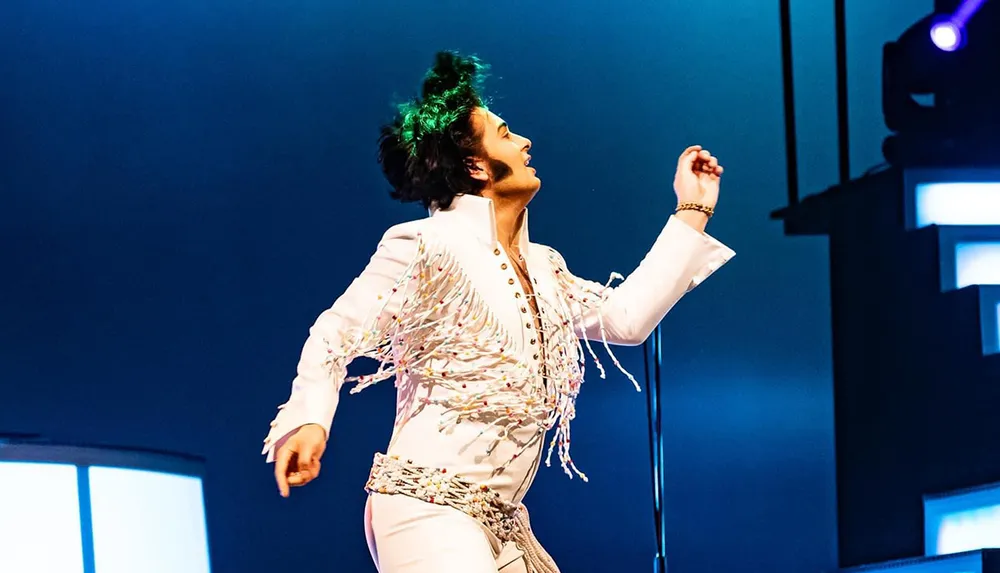 A performer wearing a white fringe jumpsuit is energetically dancing on stage