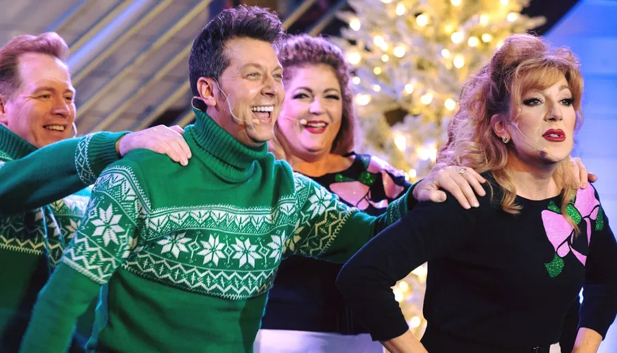 Four people in festive attire are joyfully singing or performing on a stage adorned with a Christmas tree in the background.