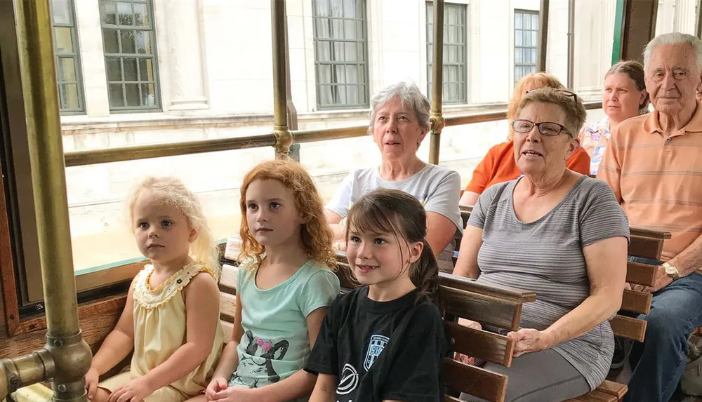 A group of people including children and seniors are seated inside a vintage-looking streetcar with some of them looking towards the camera