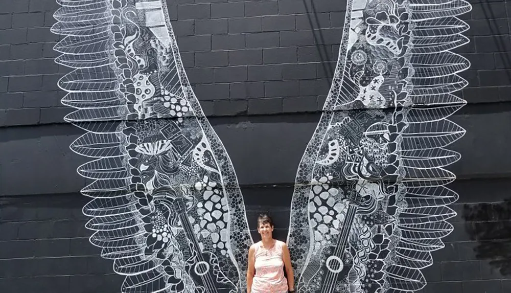 A person stands in front of a mural that resembles a large pair of intricate angelic wings creating the illusion that they are attached to their back