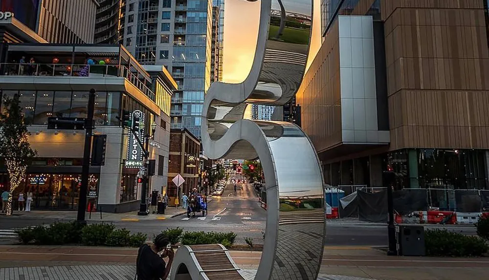 This image shows an urban scene with a large wavy metallic sculpture in the foreground overlooking a street flanked by modern buildings with people and activity visible below