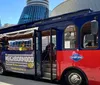 A red and blue trolley-style tour bus with passengers is parked on a city street under a clear sky