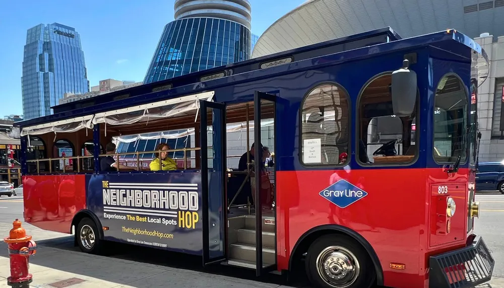 A red and blue trolley-style tour bus with passengers is parked on a city street under a clear sky