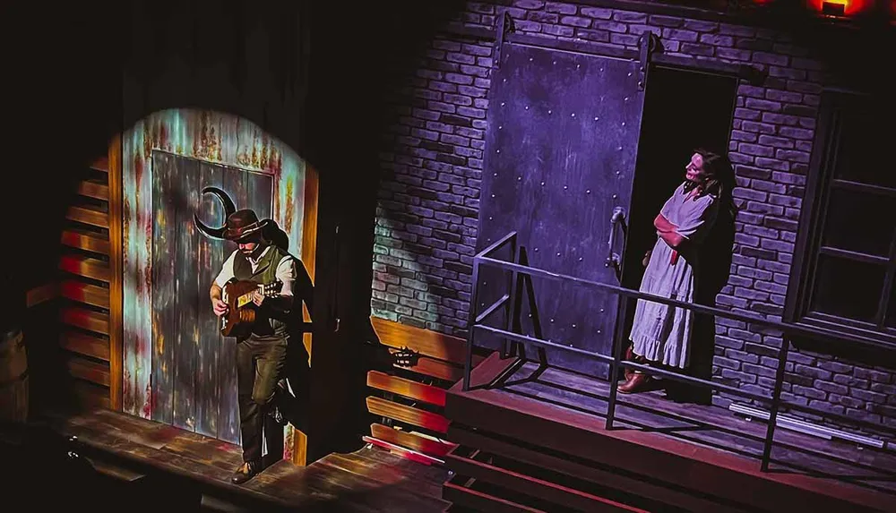 A man plays the guitar by a rustic door while a woman stands by a window on a set designed to resemble an old-timey street suggesting a scene from a theatrical production