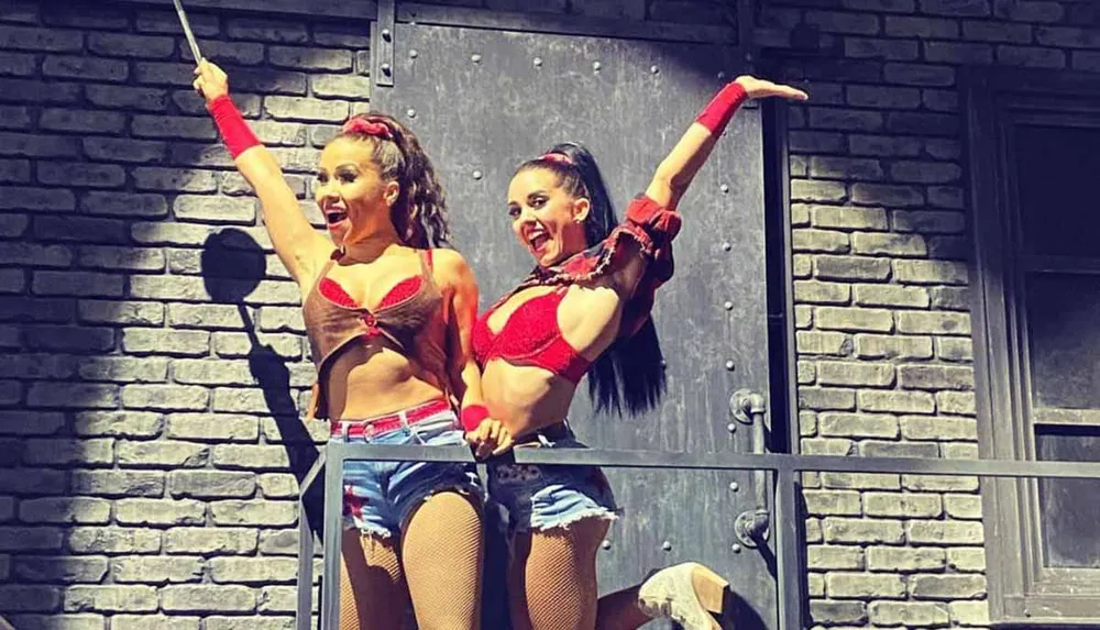 Two performers in red costumes appear to be dancing or engaging in a theatrical act on an industrial-themed stage set with a grey brick backdrop