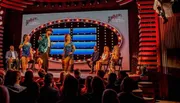 The image shows a vibrant game show set with a group of dancers performing in the foreground and the audience along with a panel of seated guests looking on.