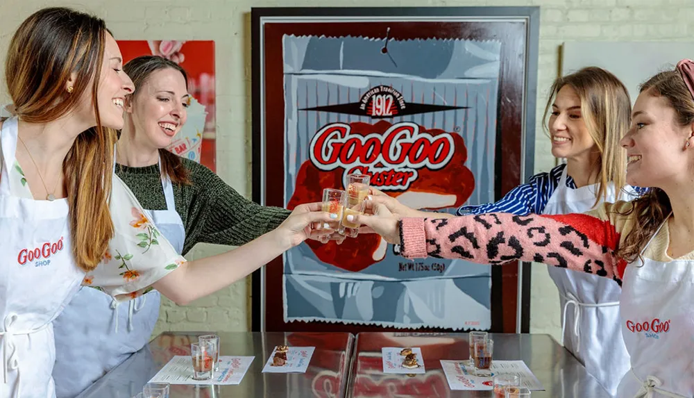 Four people wearing aprons are cheerfully toasting with small glasses in a room with a retro-styled Goo Goo Cluster advertisement poster in the background