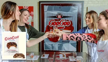 Four people wearing aprons are cheerfully toasting with small glasses in a room with a retro-styled 