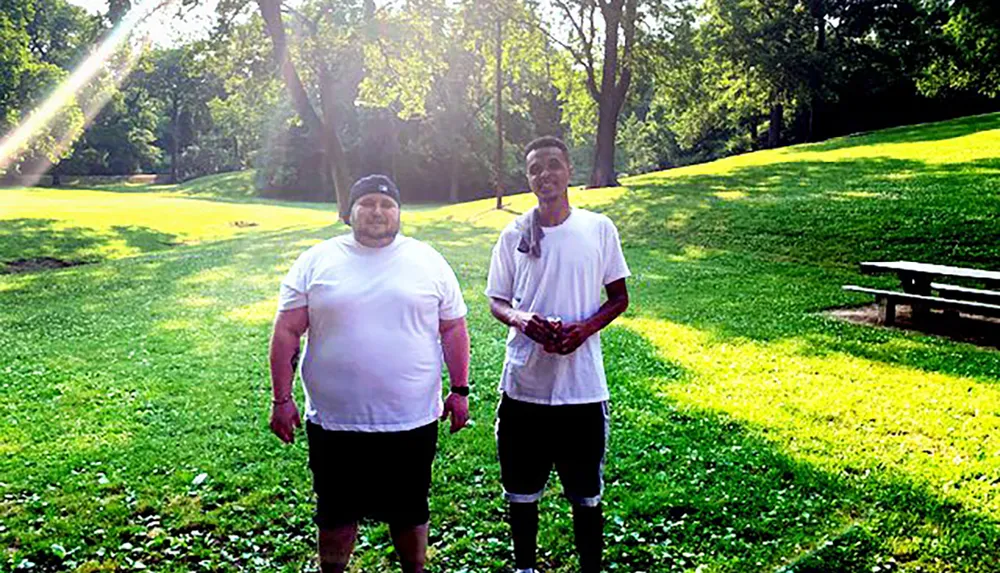 Two individuals are standing side by side in a sunlit park with trees and a bench in the background