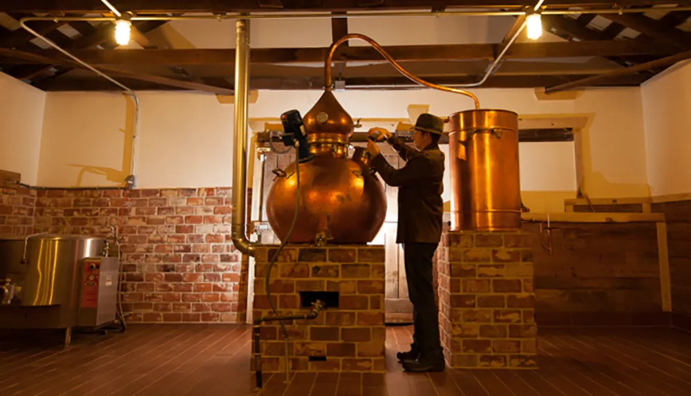 A person dressed in a suit and hat is adjusting a copper distillation apparatus in what appears to be a traditional distillery setting