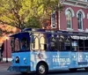 A blue trolley bus with advertising wraps for The New Franklin tour is seen on a sunny street with a backdrop of a brick building and a traffic signal showing red