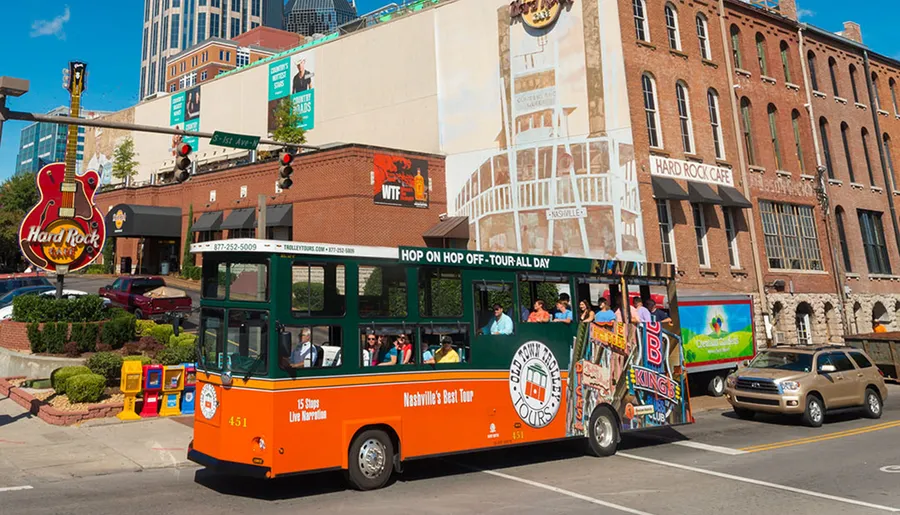 A bright orange trolley bus, labeled Nashville's Best Tour, offers a hop-on hop-off service, surrounded by city buildings including the Hard Rock Cafe, under a clear blue sky.