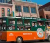 An orange and green trolley bus with the label Old Town Trolley Tours is driving past a row of historic buildings on a sunny day