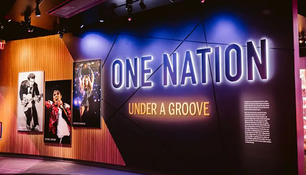 The image shows an exhibition space with the illuminated phrase ONE NATION UNDER A GROOVE and posters of iconic music artists suggesting a music-themed exhibit