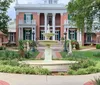 The image features an elegant pink-hued mansion with white columns and balconies flanked by two smaller structures set behind a manicured garden with a classic water fountain as the centerpiece