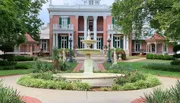 The image features an elegant pink-hued mansion with white columns and balconies, flanked by two smaller structures, set behind a manicured garden with a classic water fountain as the centerpiece.