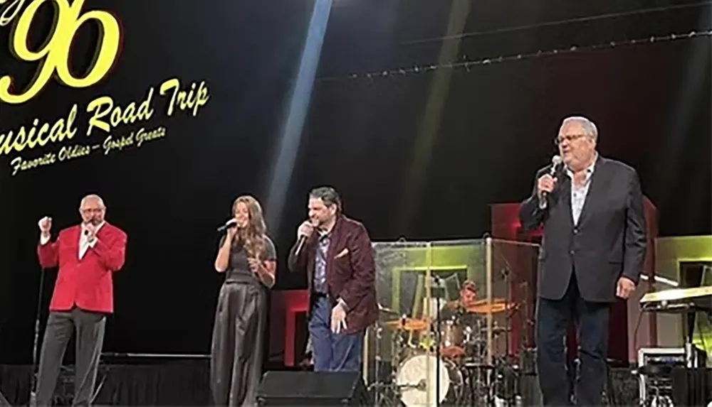 Four people are singing on stage during a musical road trip event with a drummer playing in the background