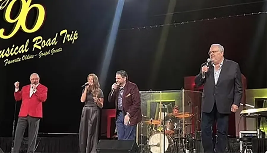 Four people are singing on stage during a musical road trip event, with a drummer playing in the background.