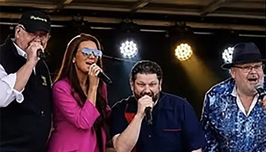 Four people are singing on stage, each holding a microphone, with lights shining behind them.