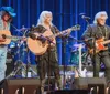 A group of six musicians perform together on stage playing acoustic guitars a banjo and a violin with a logo in the background that suggests a live music event