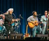 A group of six musicians perform together on stage playing acoustic guitars a banjo and a violin with a logo in the background that suggests a live music event