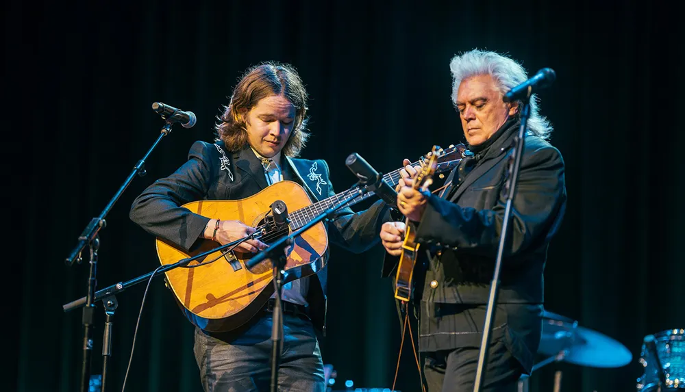 Two guitarists are intently performing on stage with one looking closely at his guitar