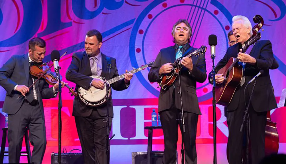 Four musicians in formal attire are performing on stage with instruments including a violin a banjo a mandolin and a guitar in front of a colorful backdrop with a musical note design
