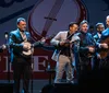 A group of six musicians perform together on stage playing acoustic guitars a banjo and a violin with a logo in the background that suggests a live music event