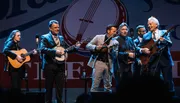 A group of six musicians perform together on stage, playing acoustic guitars, a banjo, and a violin, with a logo in the background that suggests a live music event.