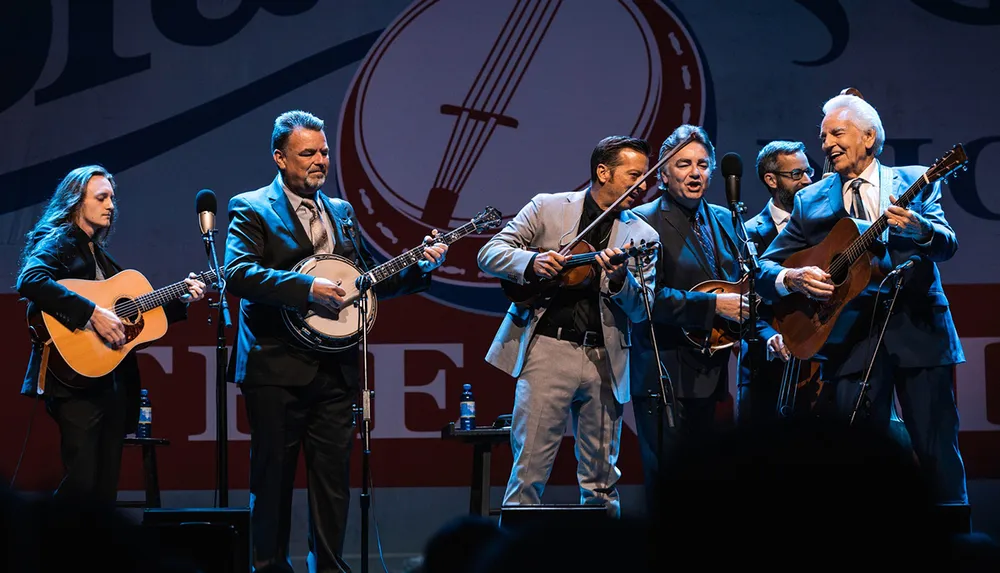 A group of six musicians perform together on stage playing acoustic guitars a banjo and a violin with a logo in the background that suggests a live music event