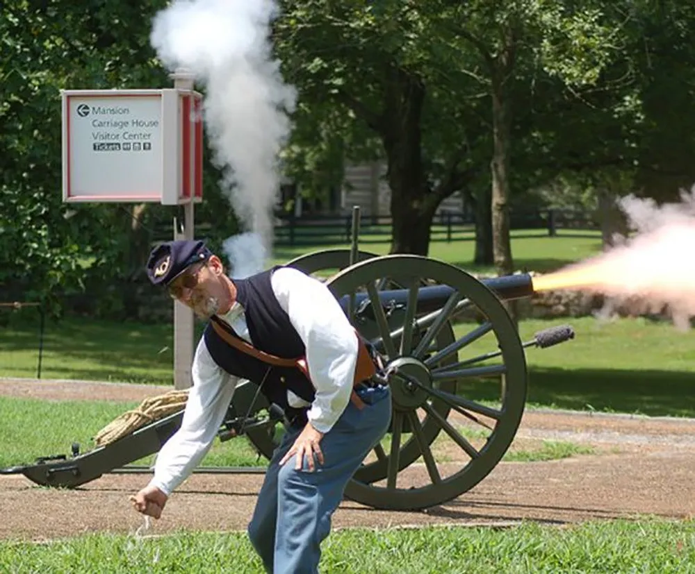 A person in historical military attire is operating a cannon that has just been fired evident by the smoke plume coming from the barrel while they are standing in a grassy area near a sign indicating a carriage house and visitor center