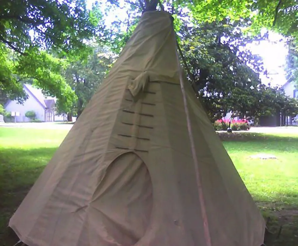 The image shows a canvas teepee tent set up on a grassy area with trees and a building in the background