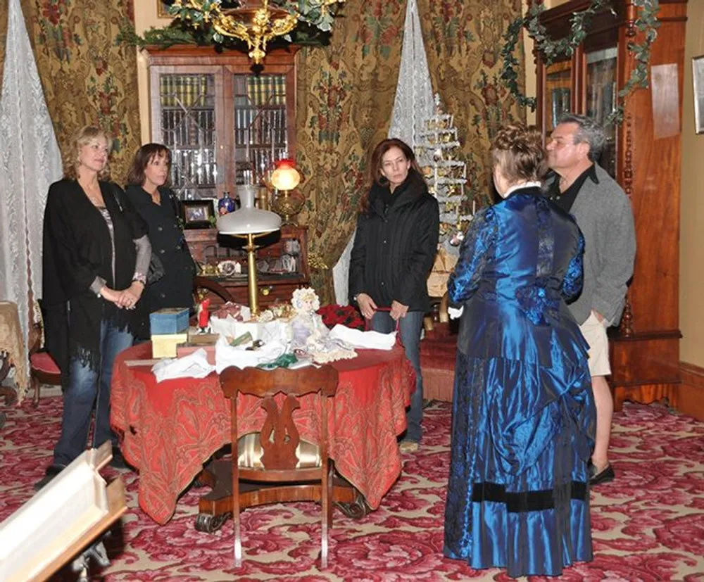 A group of people is attentively listening to a person dressed in a period costume during a tour inside a richly decorated Victorian-style room