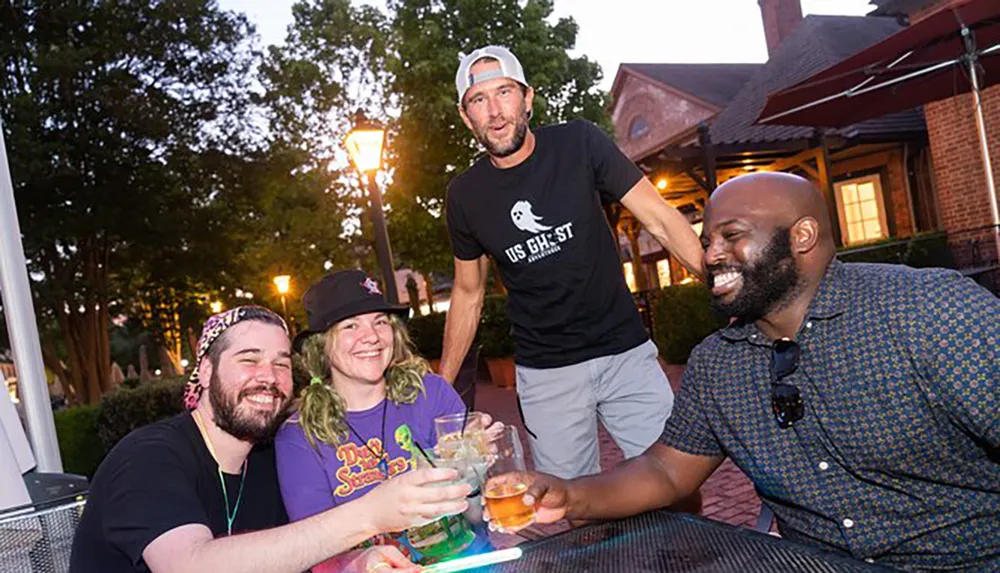 A group of four friends are sharing a light-hearted moment at an outdoor table with drinks in hand during an evening gathering