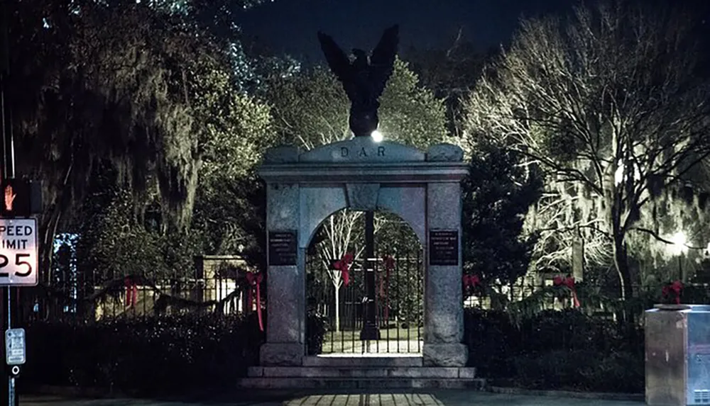 A nighttime shot of a gated monument with an eagle sculpture atop an arch surrounded by trees draped in Spanish moss
