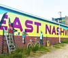 A group of people is posing for a photo in front of a large mural with vibrant artwork on a sunny day