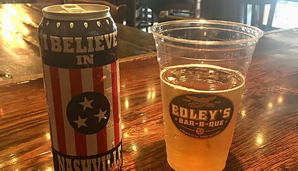 There is a patriotic-themed beer can next to a half-full plastic cup of beer on a wooden bar top with the message I BELIEVE IN NASHVILLE inscribed on the can