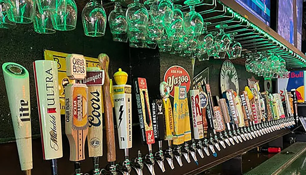 The image shows a variety of beer tap handles lined up on a bar with an array of hanging glasses above them