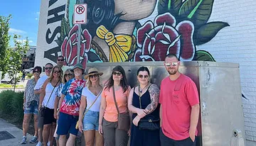 A group of people is posing for a photo in front of a large mural with vibrant artwork on a sunny day.