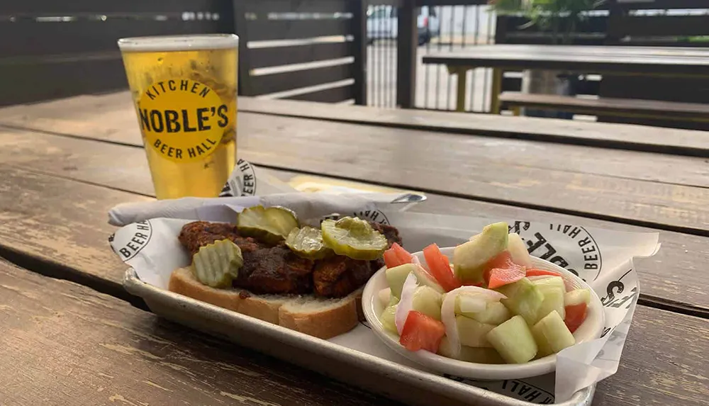On a wooden picnic table there is a pint of beer next to a tray containing a sandwich with pickles and a side of chopped vegetables