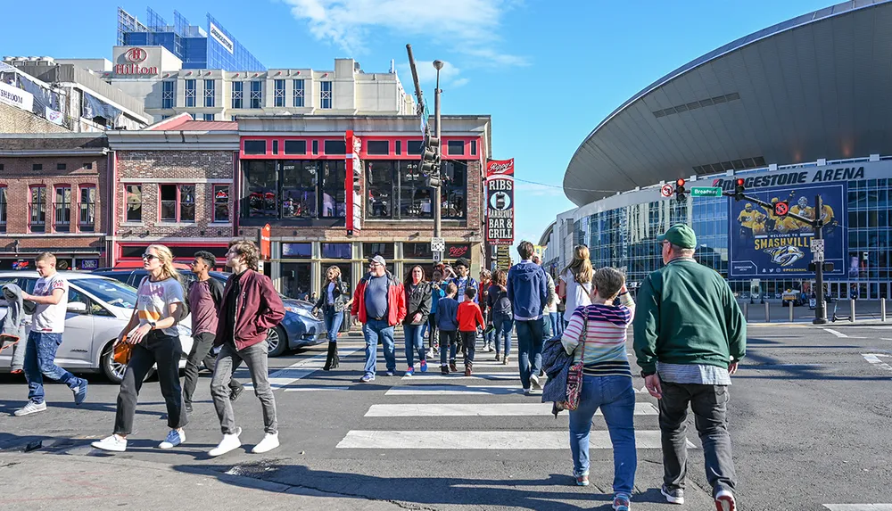 People are walking across a zebra crossing on a sunny day with the Bridgestone Arena in Nashville Tennessee visible in the background