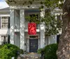 The image shows a two-story white house with black shutters a red entrance door and a red banner hanging from the second-floor balcony all framed by green foliage and a prominent tree