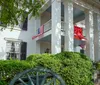 The image shows a two-story white house with black shutters a red entrance door and a red banner hanging from the second-floor balcony all framed by green foliage and a prominent tree