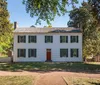 The image features a two-story white colonial house with green shutters and a red door surrounded by trees and a well-kept lawn under a clear blue sky