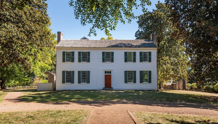 The image features a two-story white colonial house with green shutters and a red door, surrounded by trees and a well-kept lawn under a clear blue sky.