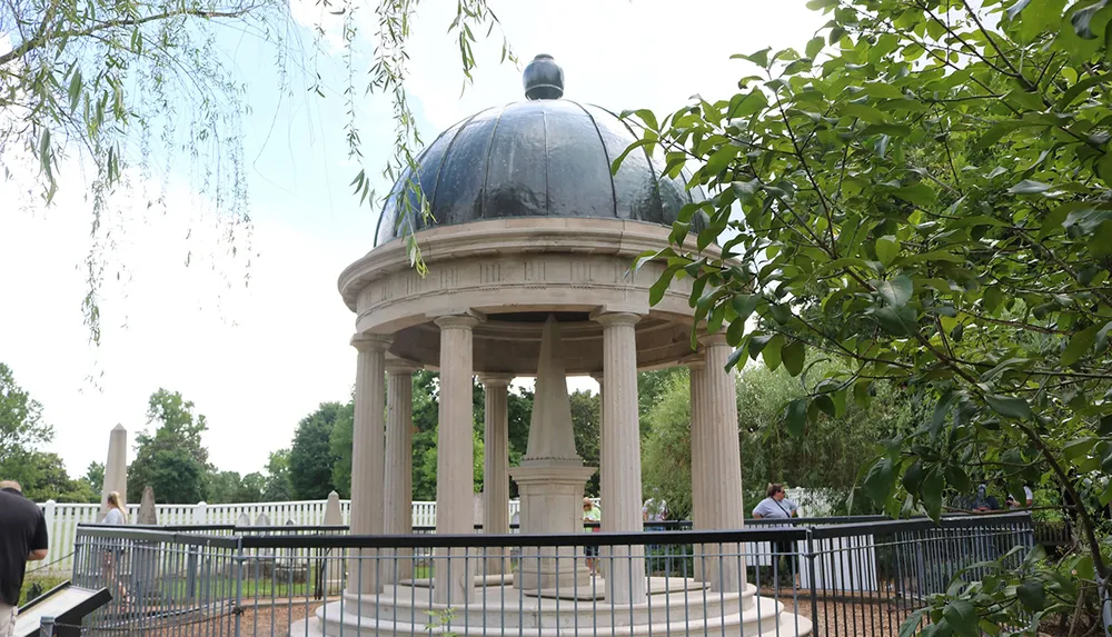 The image shows a classical-style gazebo with a dark dome top surrounded by greenery and visitors enclosed by a low metal fence
