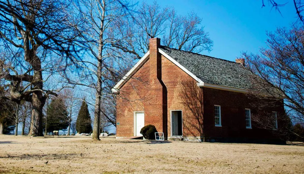 A quaint single-story red brick house with a gabled roof sits on a lawn with trees under a clear blue sky