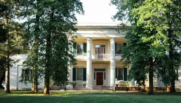The image shows a stately two-story house with white columns and shutters, surrounded by mature trees and a well-kept lawn.