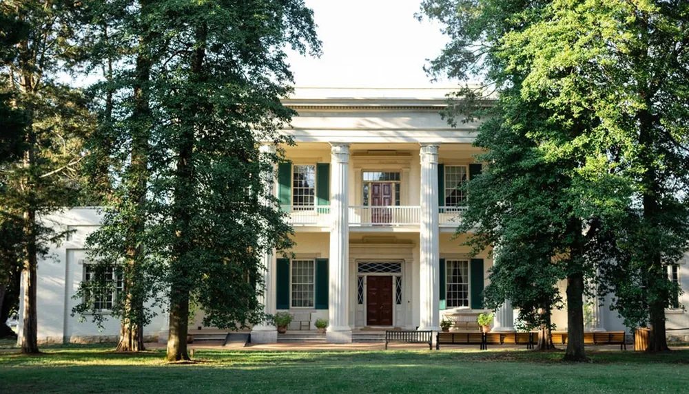 The image shows a stately two-story house with white columns and shutters surrounded by mature trees and a well-kept lawn